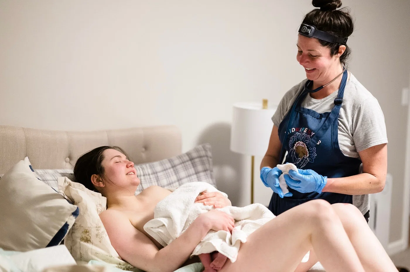 A midwife smiling beside a mother resting with her newborn immediately after a home birth